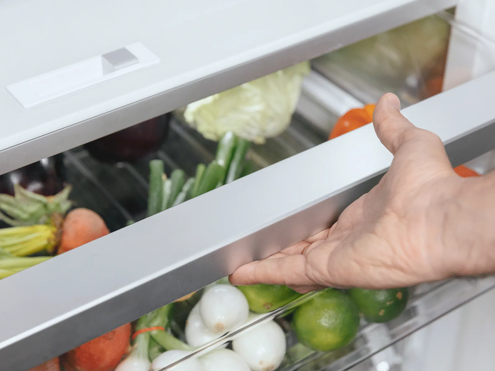 Hand pulling open a refrigerator crisper drawer filled with assorted fresh vegetables — carrots, limes, small white onions, and leafy greens visible through