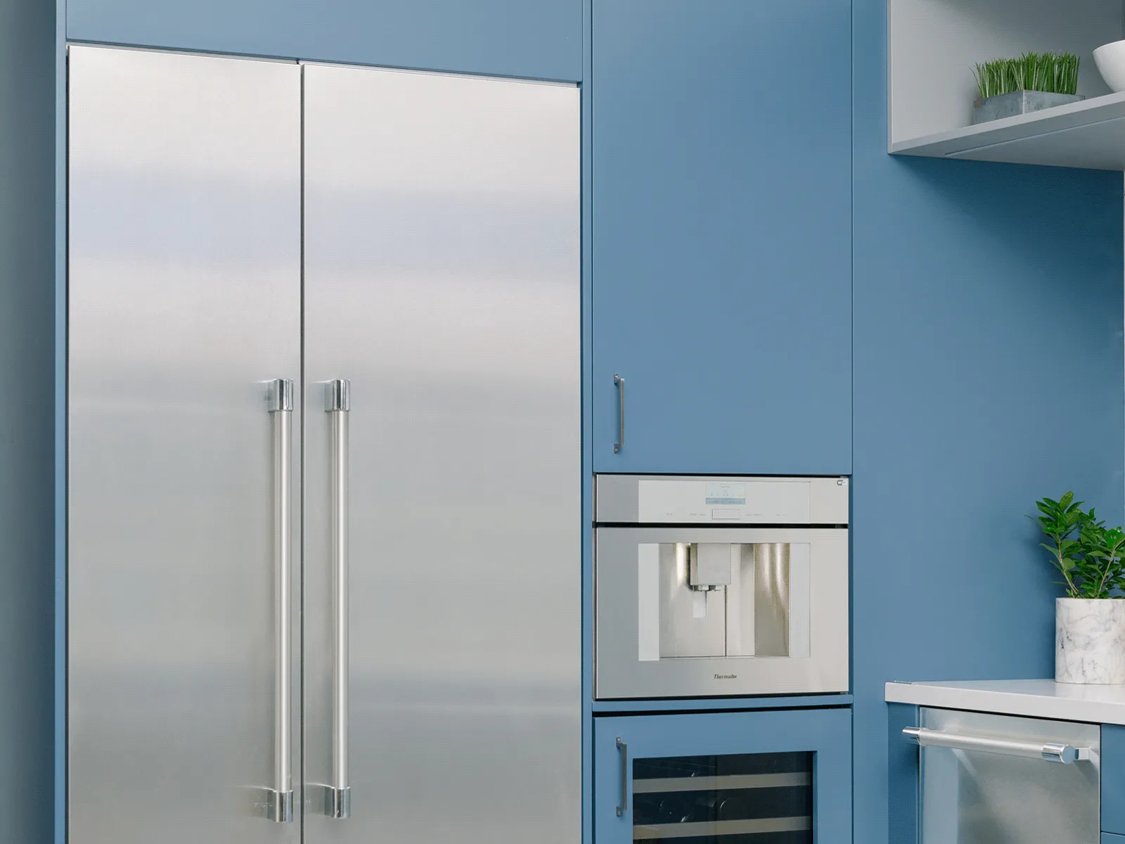 Contemporary kitchen scene with blue cabinetry: a stainless-steel side-by-side refrigerator with long vertical handles built into the blue cabinets, a built-in coffee dispenser above a glass-front wine cooler, a stainless-steel dishwasher to the right, a white countertop with a potted plant and cutting board, open shelving holding dishes, and gray tile floor.