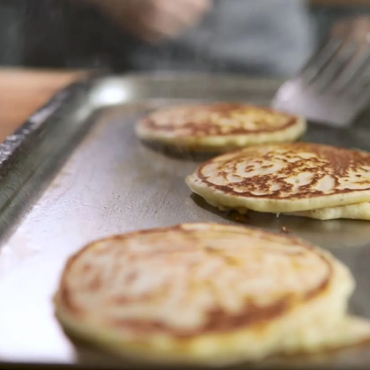 Three golden-brown pancakes are cooking on a large silver griddle on an induction cooktop, with a spatula flipping one.