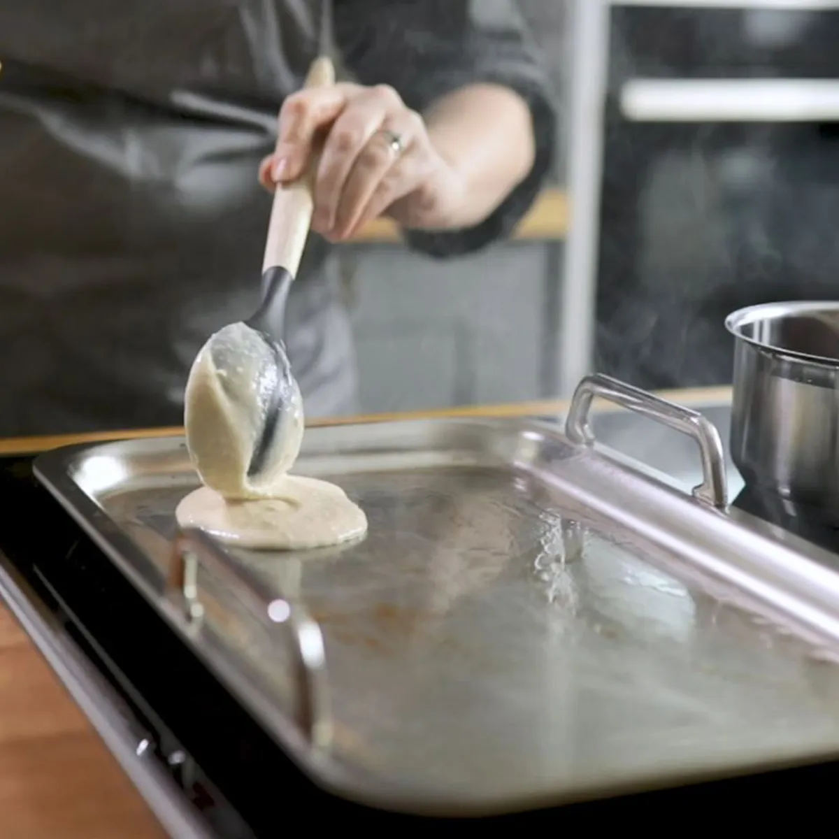 A hand uses a ladle to pour pancake batter onto a large silver griddle on an induction cooktop, with a saucepan of sauce simmering nearby.