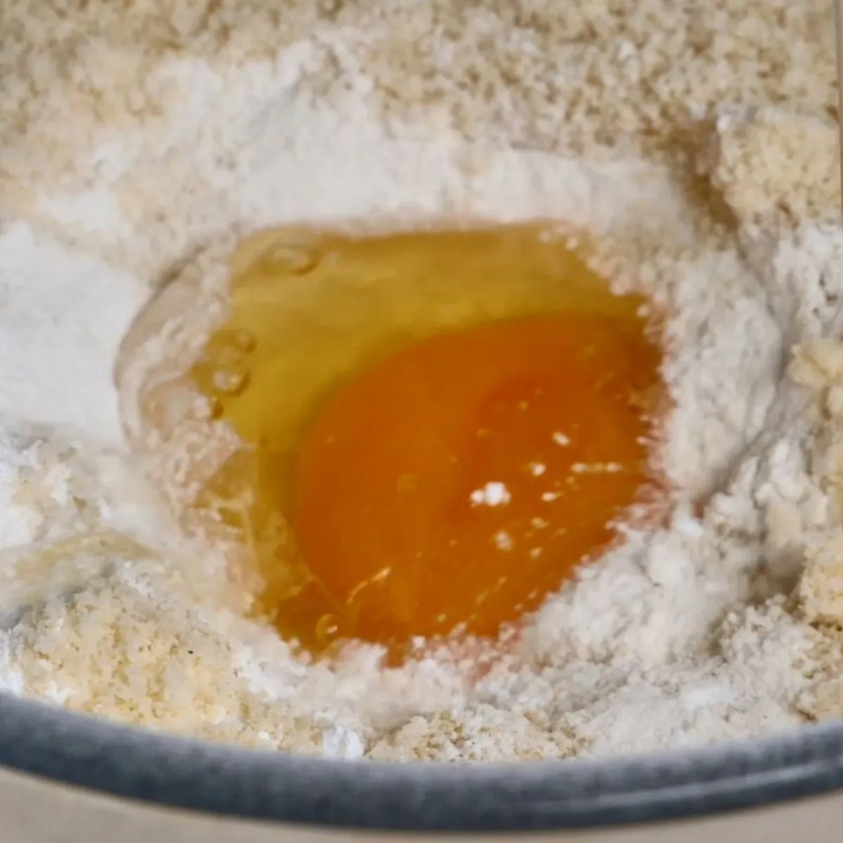 A close-up of a glass bowl containing white flour and almond flour, with a cracked egg and a drizzle of oil in the center.