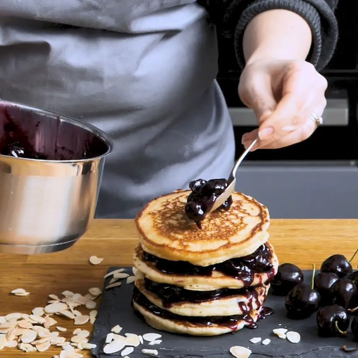 A hand spoons dark cherry sauce from a saucepan onto a stack of golden pancakes, garnished with fresh cherries and almond flakes, on a black slate board.