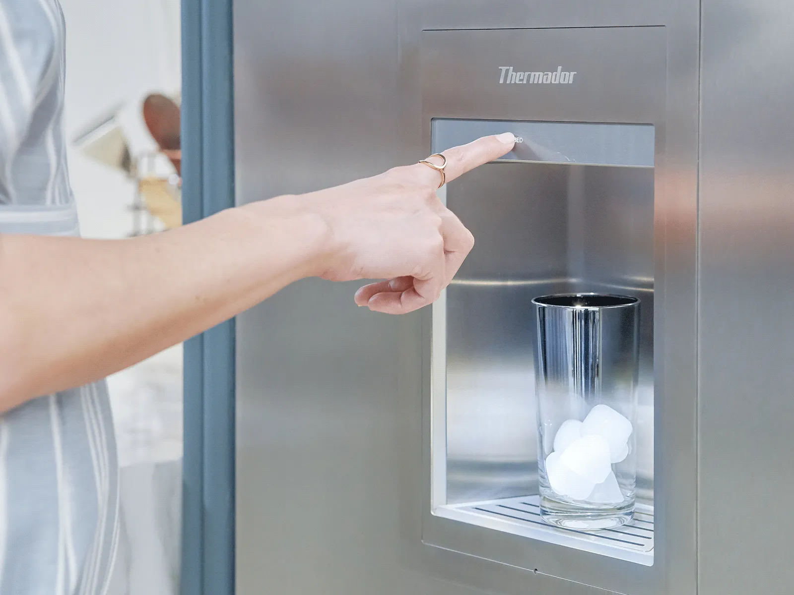 Hand pressing button on a stainless steel refrigerator ice dispenser, dispensing ice cubes into a glass.