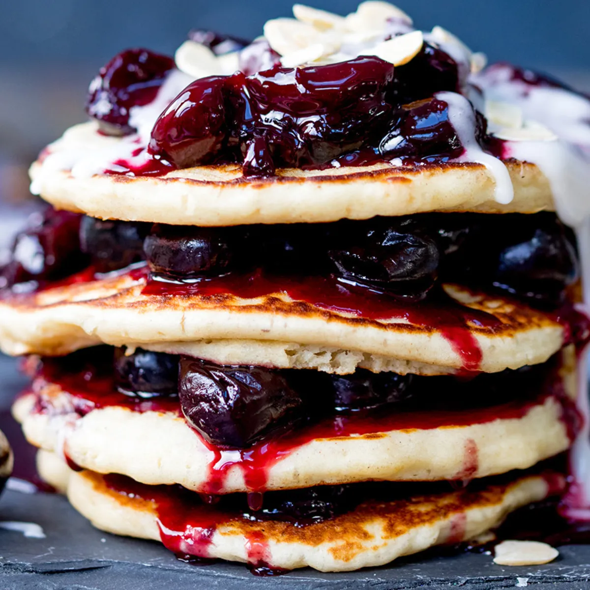 A stack of four pancakes topped with dark cherry sauce, white cream, and almond flakes, surrounded by fresh cherries and a jar of cherry sauce, on a black slate board.