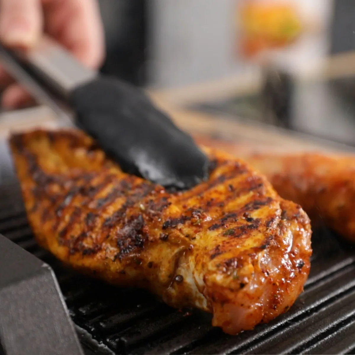 A hand uses tongs to flip a grilled chicken breast on a hot grill pan, showing prominent grill marks, next to a sausage.