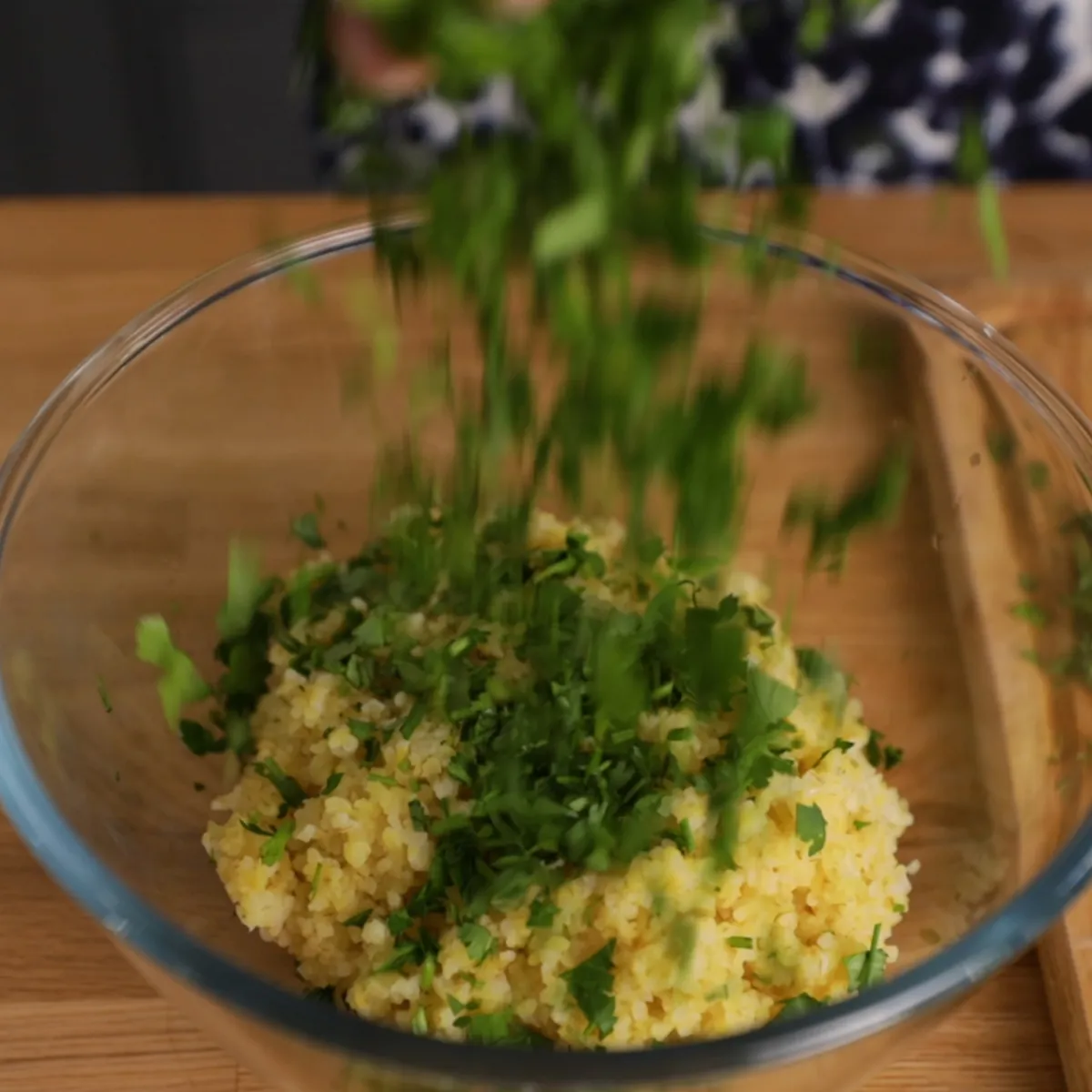 Chopped fresh parsley is sprinkled into a clear glass bowl containing cooked couscous.