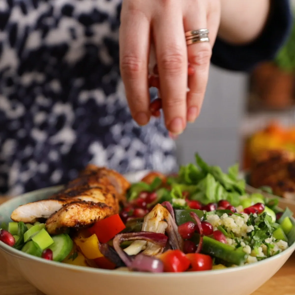 A hand sprinkles pomegranate seeds over a colorful salad bowl with grilled chicken, roasted vegetables, and couscous.
