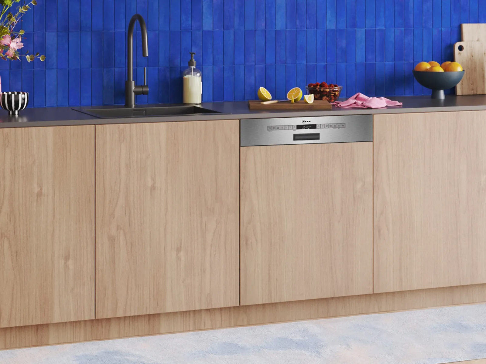 Modern kitchen with wooden cabinetry, a black countertop, blue tiled backsplash, and colorful decor, including flowers and fruit.
