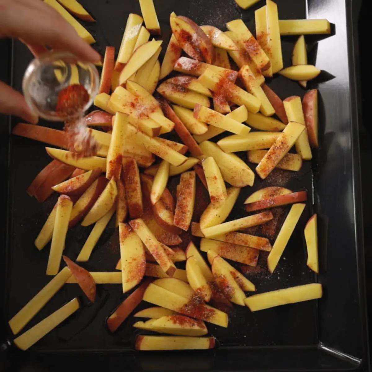 A hand sprinkles red seasoning from a small glass shaker over a baking tray filled with raw potato fries, next to grilled flatbreads.