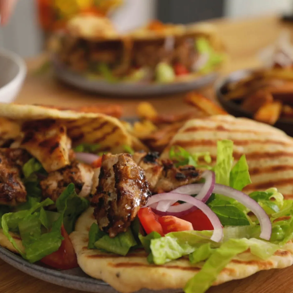 A hand uses tongs to add grilled chicken pieces to a plate with grilled flatbread, lettuce, red onion, and tomatoes, assembling a gyro.