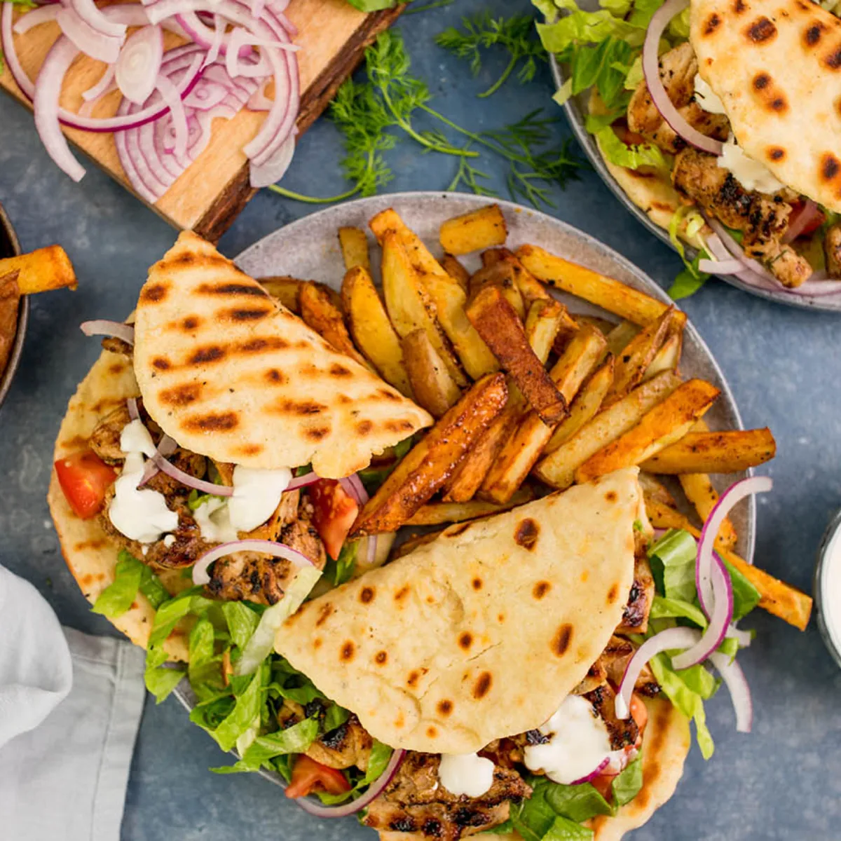 An overhead view of two plates of chicken gyros with grilled flatbread, lettuce, red onion, tomatoes, and a white sauce, served with a side of seasoned fries and a bowl of white sauce.