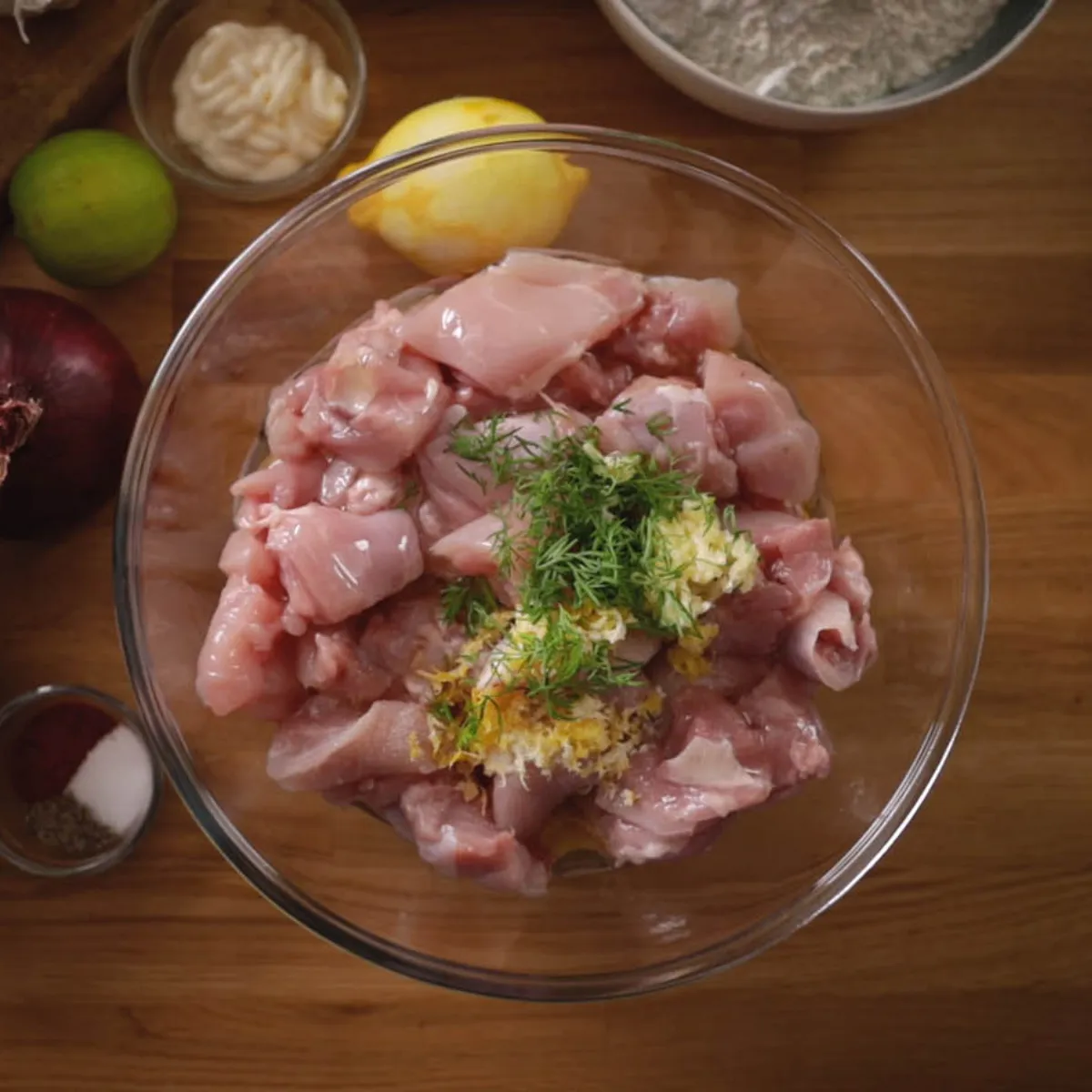 An overhead view of a clear glass bowl filled with diced raw chicken, minced garlic, lemon zest, and fresh dill, surrounded by various ingredients like tomatoes, onions, and spices on a wooden countertop.