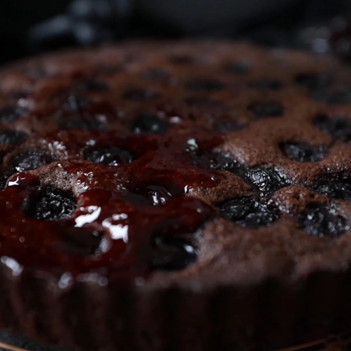 A hand uses a black pastry brush to glaze a baked chocolate and cherry tart with a shiny red sauce on a wire rack.
