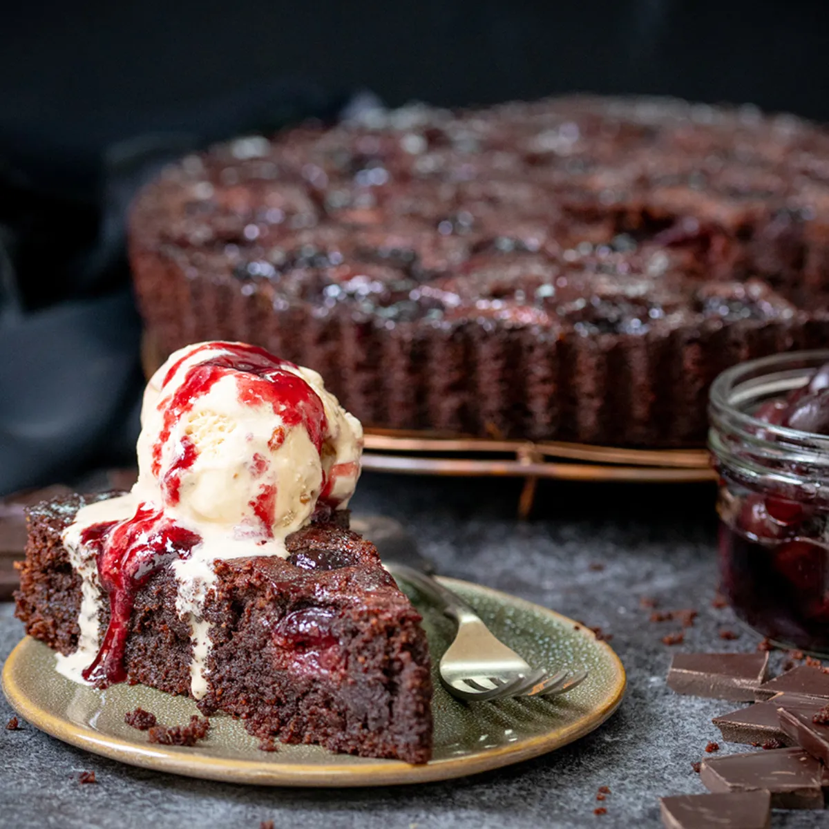 A slice of rich chocolate and cherry tart topped with vanilla ice cream and cherry sauce, served on a green plate next to the whole tart, a jar of cherries, and chocolate pieces.
