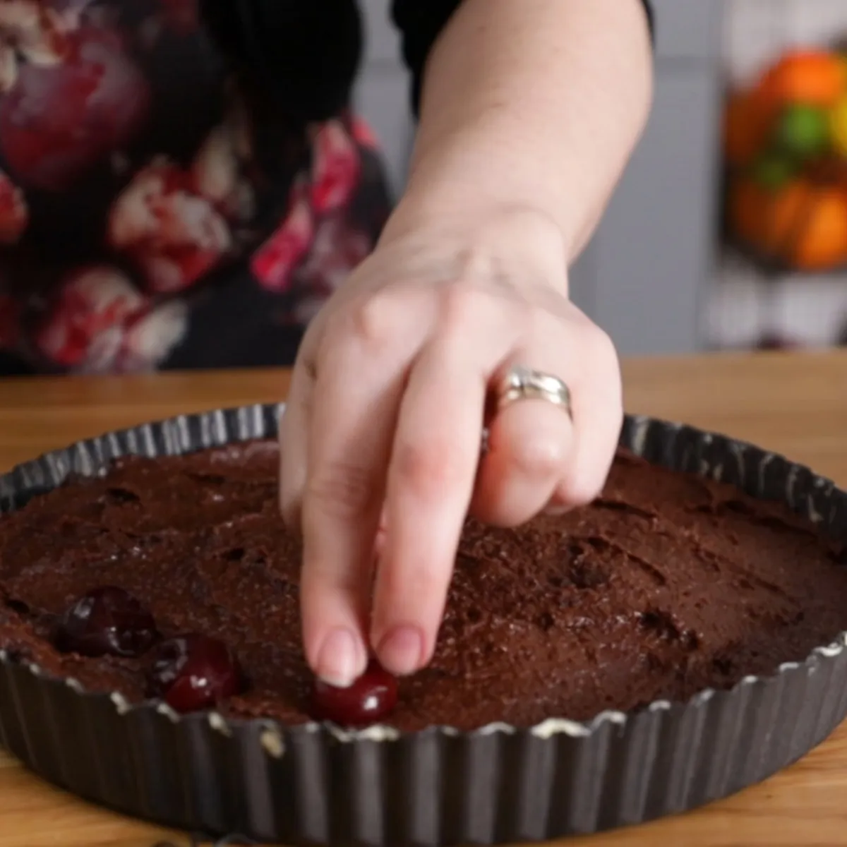 A hand places fresh red cherries onto a dark chocolate tart batter in a fluted tart pan.