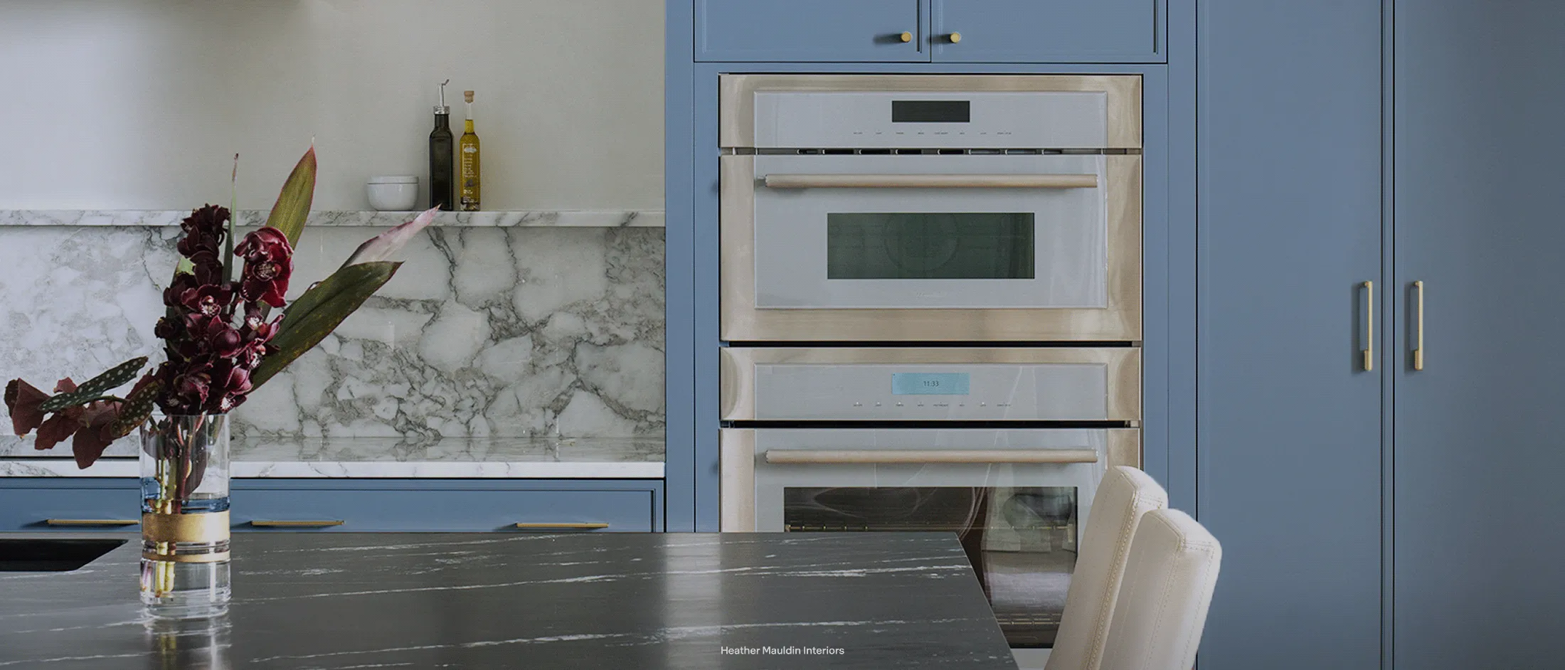 Modern kitchen with blue cabinets, built-in double oven, marble backsplash, and a vase with dark red flowers on the counter.