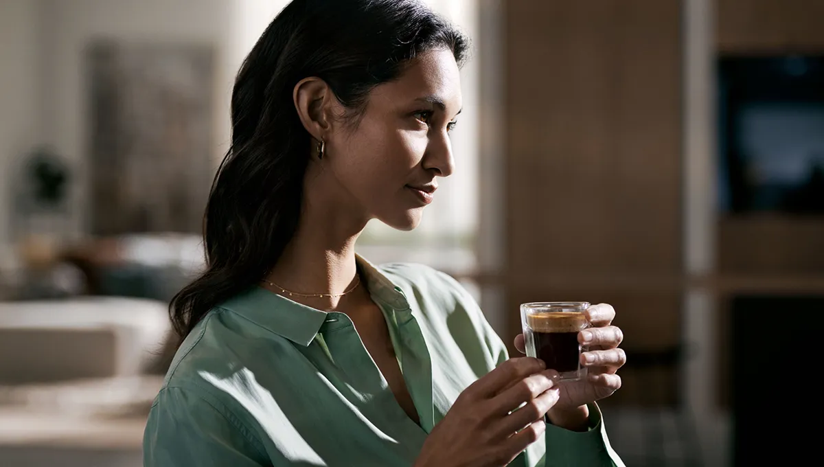 Woman in green shirt holding glass of dark liquid, sitting indoors.