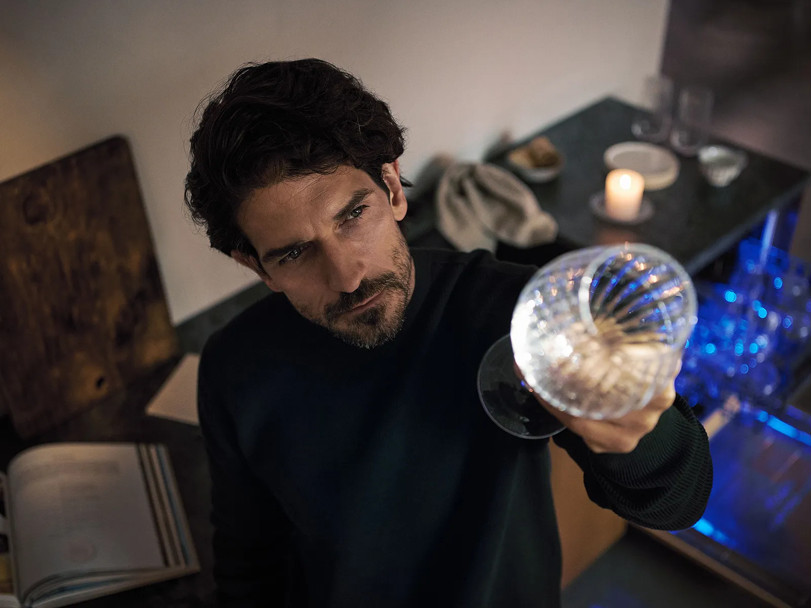 Person holding a glass with a beverage, standing near a counter with a lit candle, glasses, and cloth in a dimly lit setting.