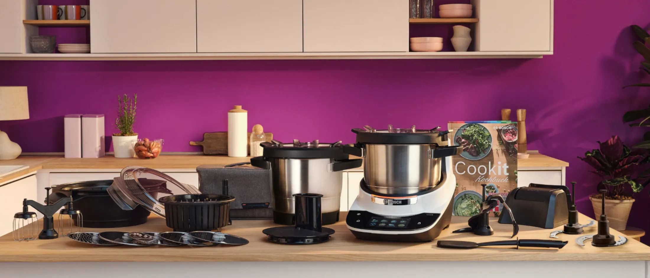 Plan de travail de cuisine avec l'appareil de cuisson Bosch Cookit, plusieurs accessoires de bol, des lames de découpe, divers accessoires et un livre de recettes Cookit.