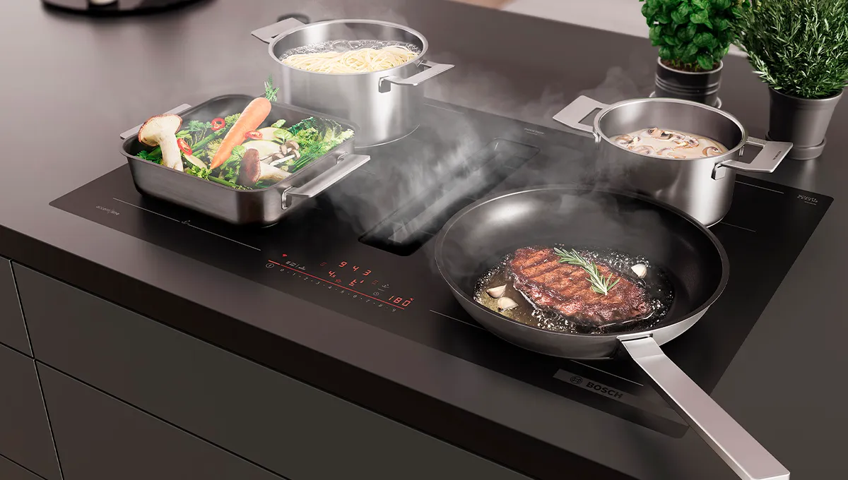 Encimera de inducción Bosch con extractor integrado, cocinando filete, verduras, pasta y champiñones. El vapor se eleva hacia la ventilación.