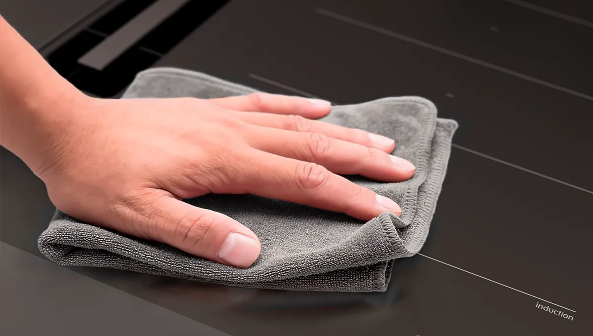 Mano limpiando una encimera de inducción negra con un paño de microfibra gris.