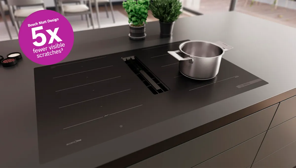 Elegante encimera de inducción Bosch negra con una olla humeante, ventilación integrada y dos pequeñas plantas en una moderna isla de cocina.
