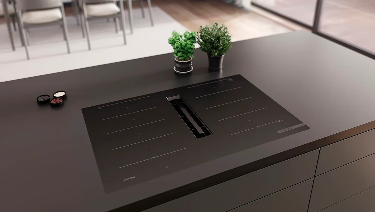 Elegante encimera de inducción Bosch negra, ventilación integrada y dos pequeñas plantas en una moderna isla de cocina.