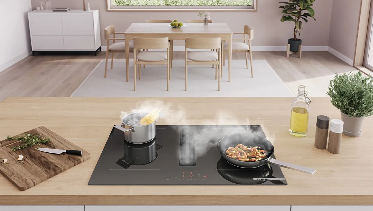 Cocina moderna con una encimera en isla donde se hierve pasta y se saltean verduras, zona de comedor al fondo y plantas en la encimera.