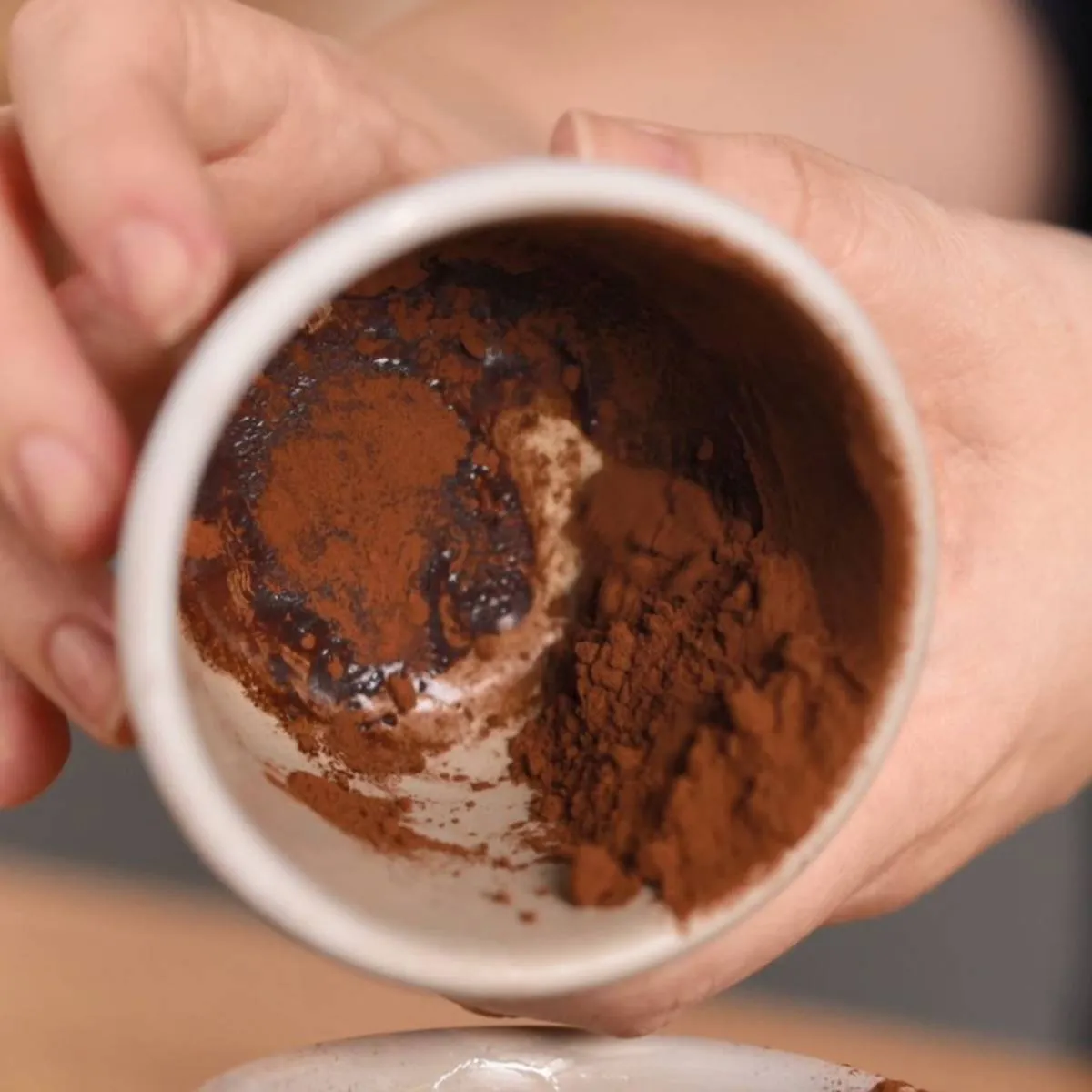 Hands hold a small white cup, tilted to show a mixture of dark cocoa powder and a dark liquid (possibly coffee or melted chocolate) inside.