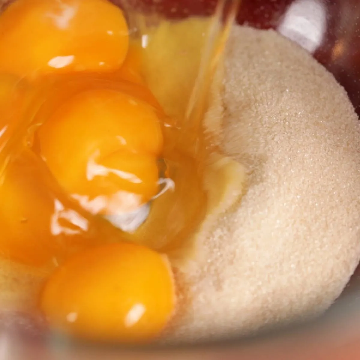 A close-up of a clear glass mixing bowl containing several egg yolks and white granulated sugar.
