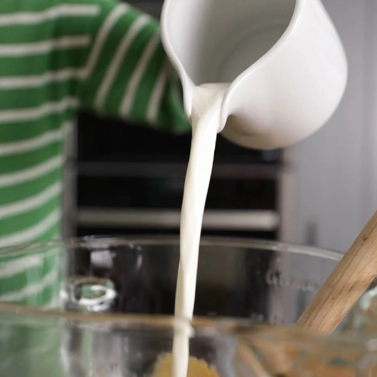 A hand pours white liquid (likely milk or cream) from a white pitcher into a large clear glass mixing bowl.