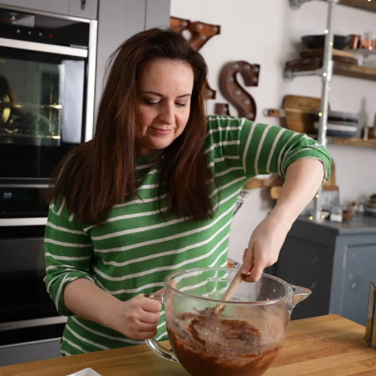 A woman in a green and white striped sweater mixes dark chocolate cake batter in a large clear glass bowl with a wooden spoon in a kitchen.