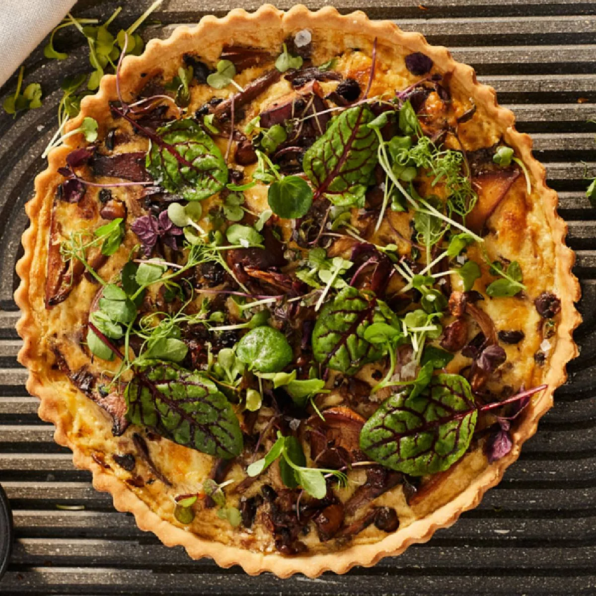 Savory quiche with greens, herbs, and a flaky crust on a dark baking tray.