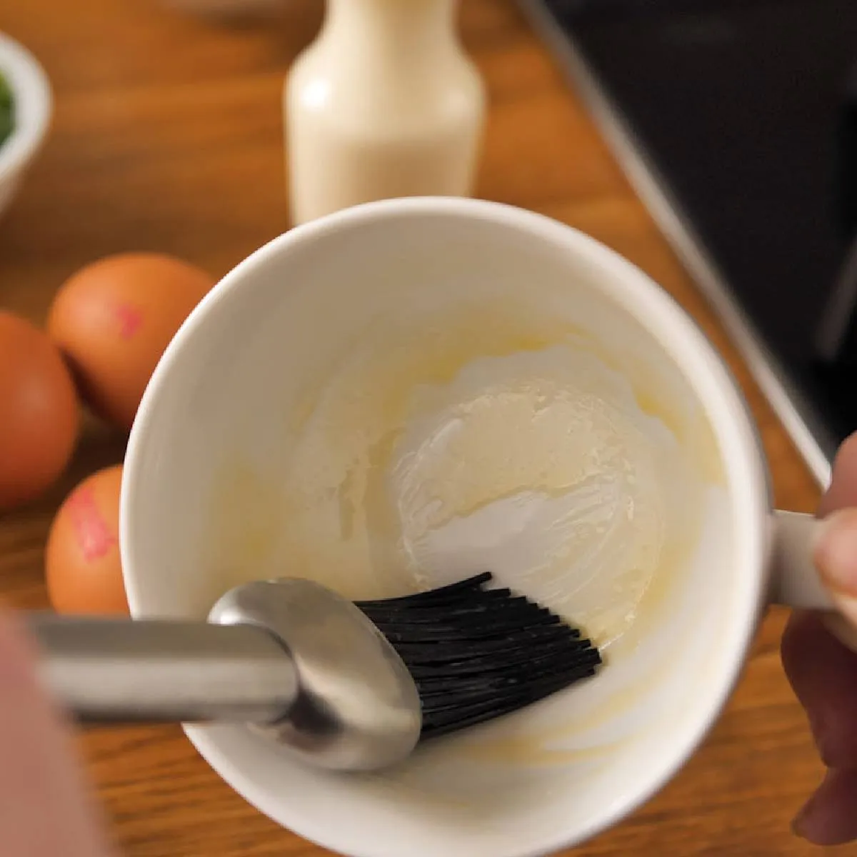 Hands use a black pastry brush to grease the inside of a white mug with oil.