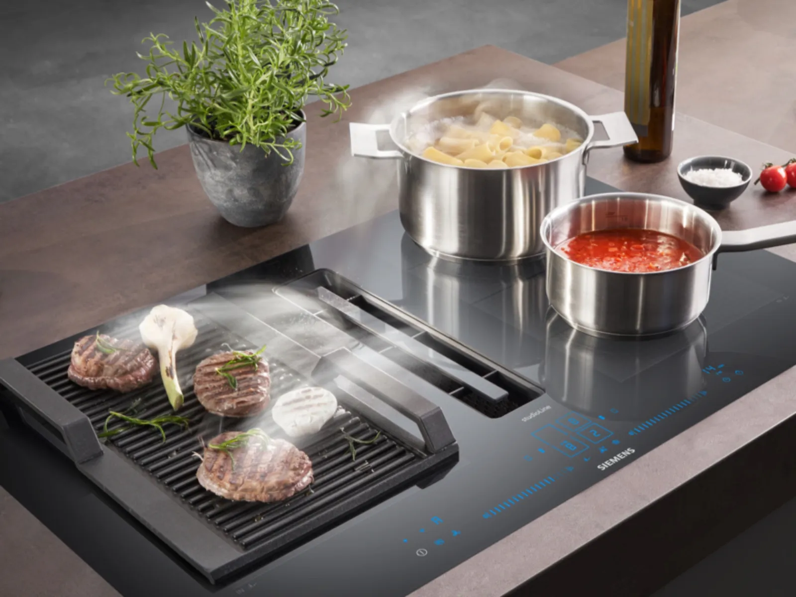Pentole in acciaio inox, carne alla griglia, erbe aromatiche e un bicchiere di vino rosso su un moderno piano di lavoro da cucina.