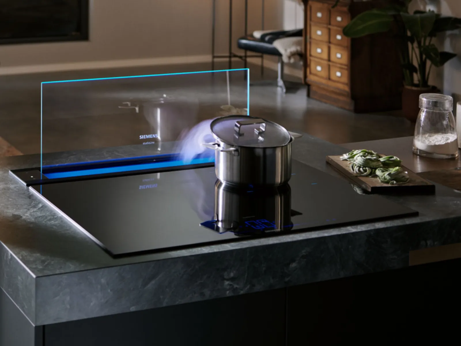 Una moderna isola cucina con un elegante piano cottura a induzione nero, una pentola in acciaio inox da cui fuoriesce del vapore e una cappa da piano a scomparsa Siemens.