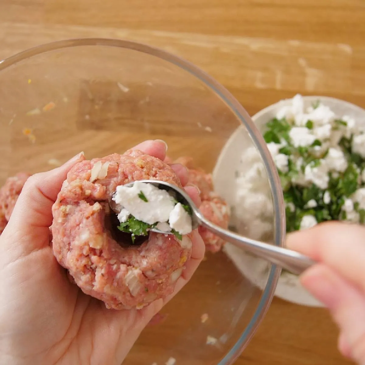 Hands use a spoon to fill the center of a raw burger patty with white crumbled feta cheese and green herbs.