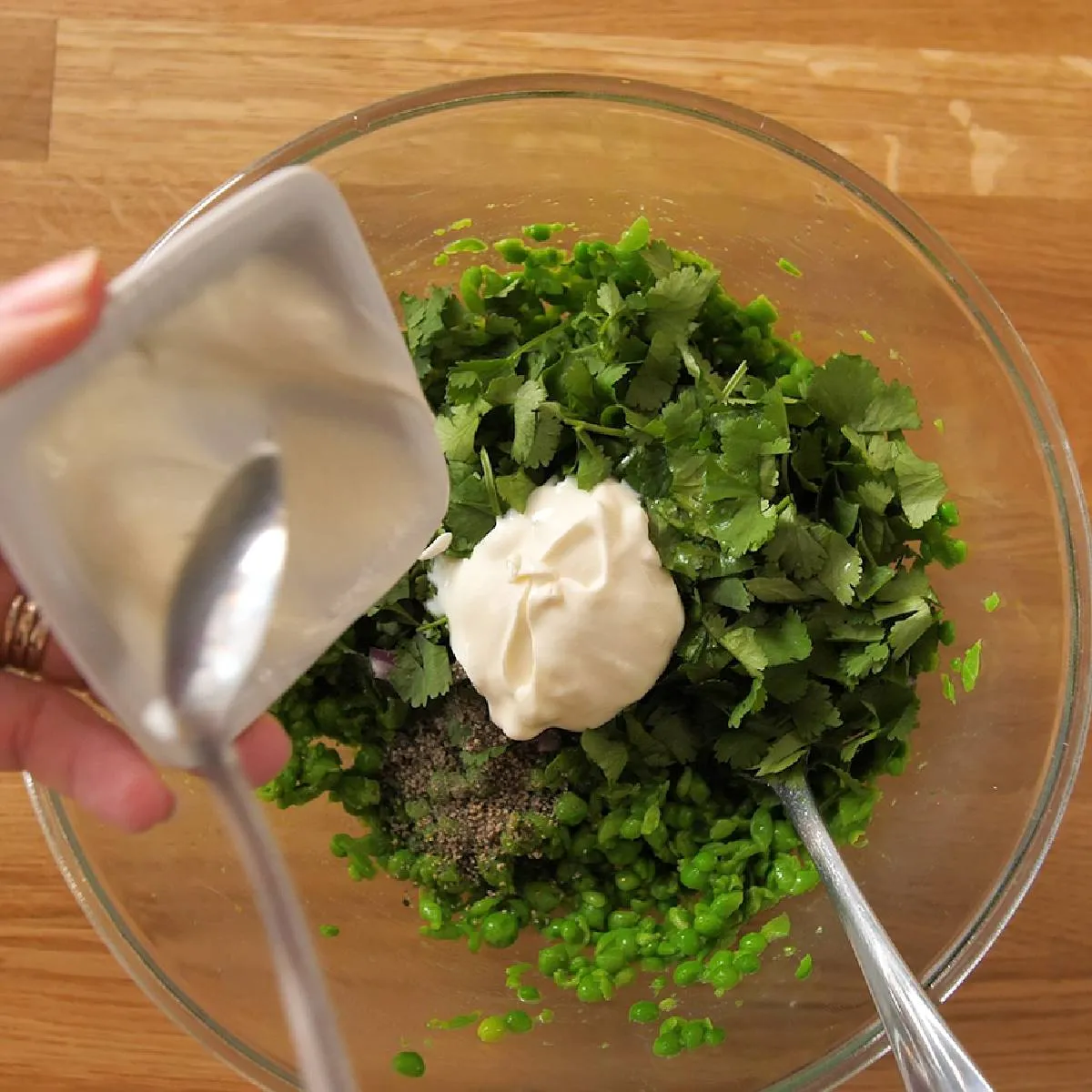 An overhead view of a hand adding a spoonful of white dressing to a glass bowl containing mashed green peas and chopped cilantro.