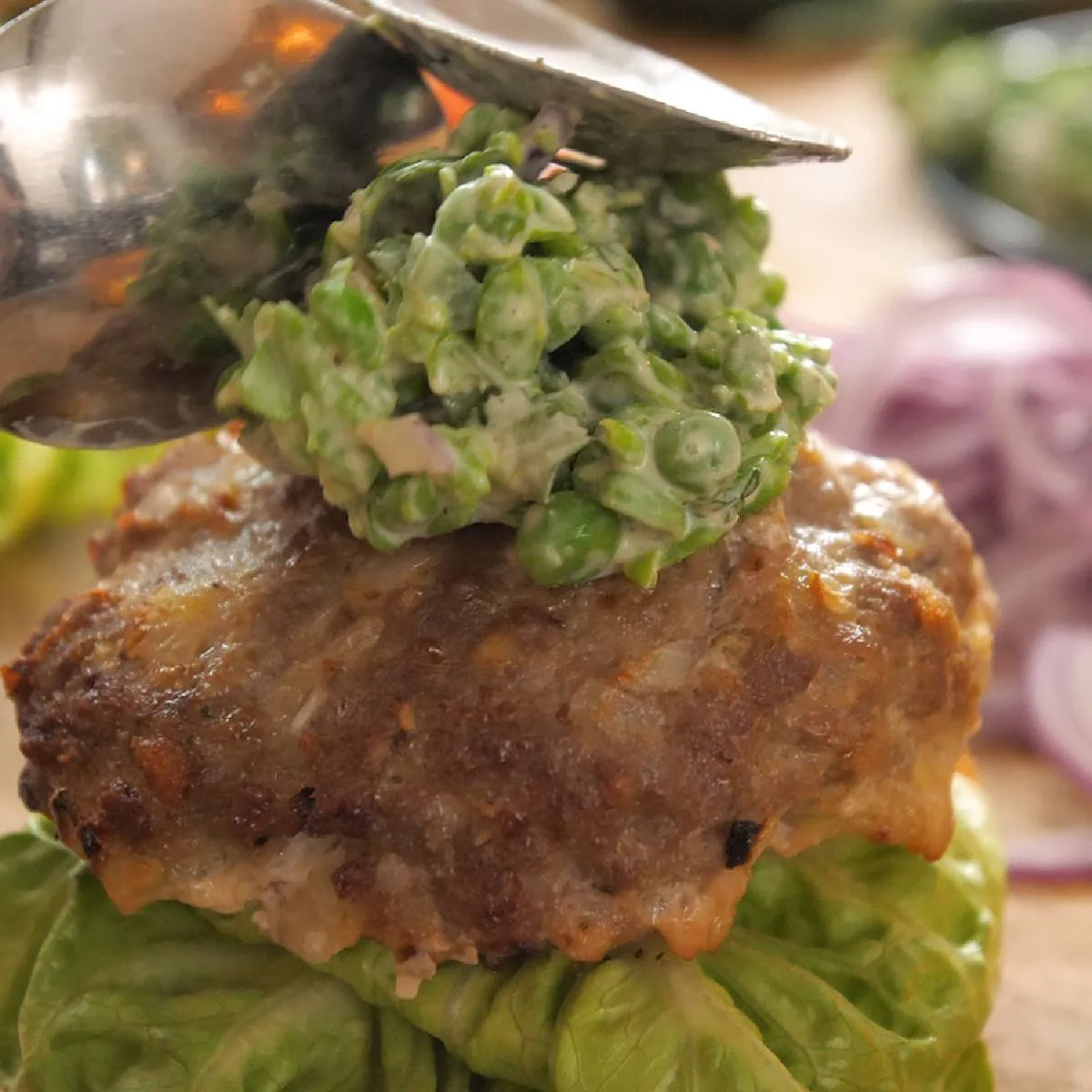 A hand uses a spoon to dollop a green pea and feta mash onto a cooked burger patty, which rests on a bed of lettuce.