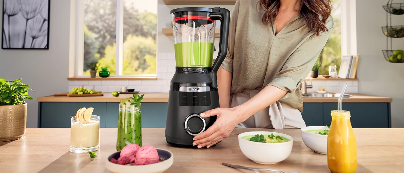 Una persona utilizza un frullatore Bosch High Performance nero VitaPower Series 6 riempito con frullato verde in cucina. Sul piano di lavoro sono disposti vari frullati e ciotole.