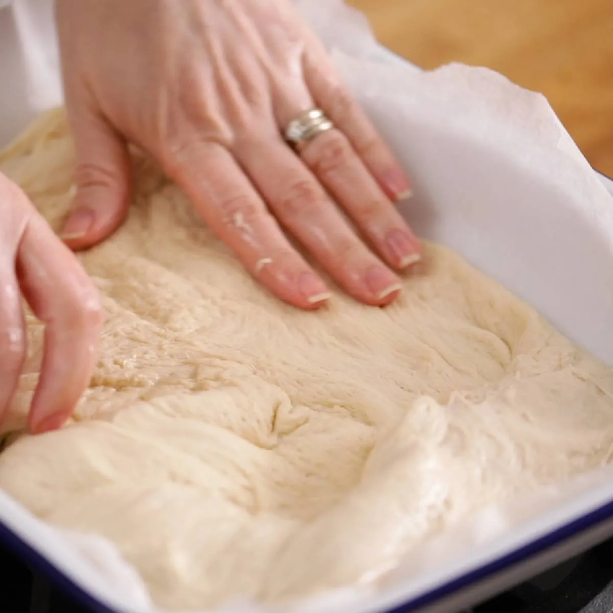 Hands gently press and spread a raw focaccia dough into a white and blue enamel baking dish lined with parchment paper.