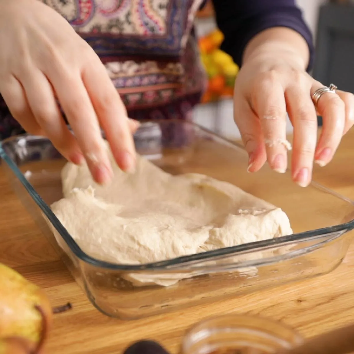 Hands transfer a large piece of raw focaccia dough into a clear glass baking dish.