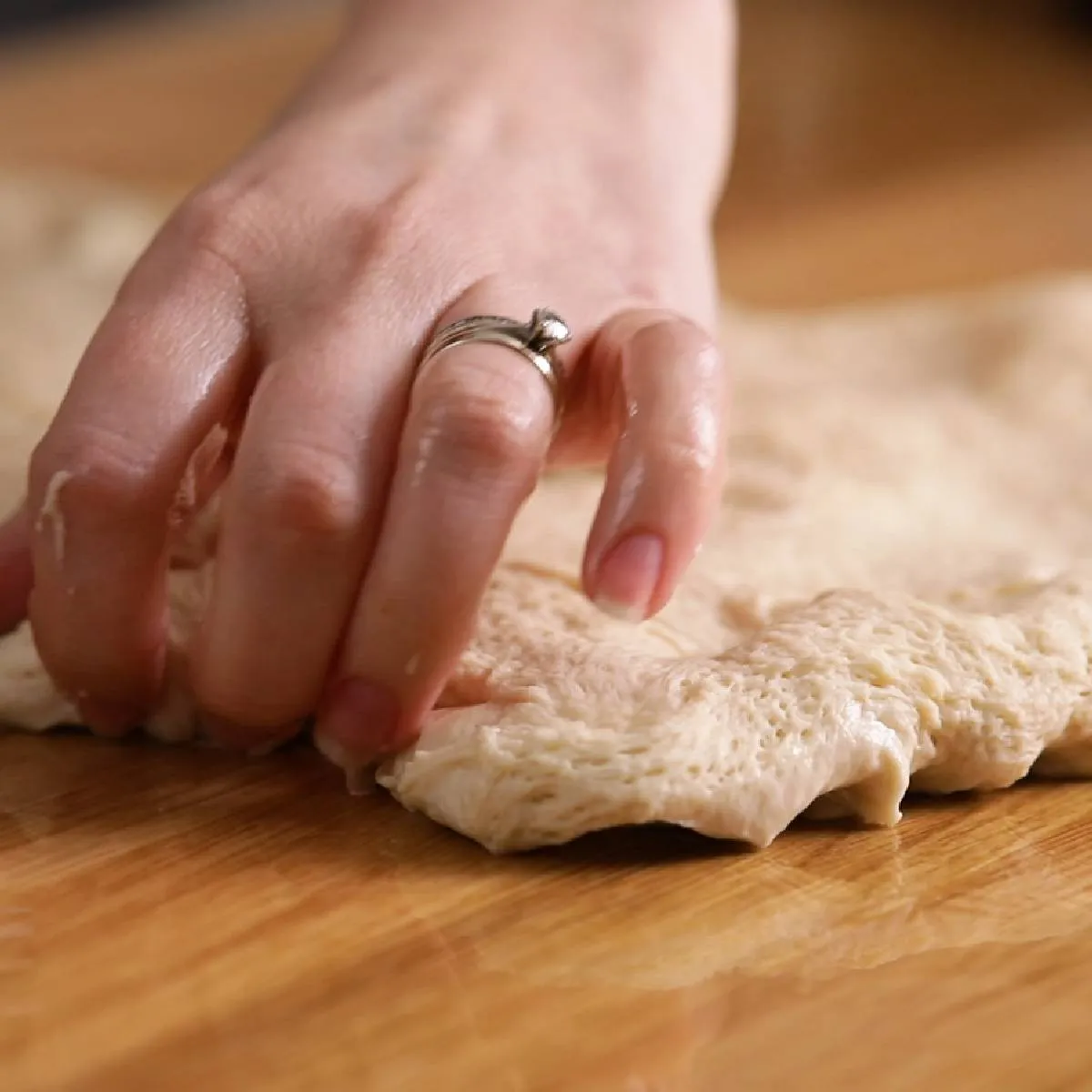 A hand gently stretches and folds a piece of raw focaccia dough on a wooden surface.