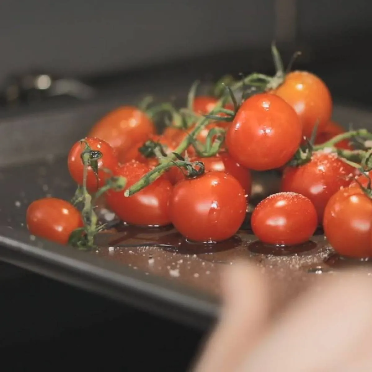 A hand slides a baking tray of fresh cherry tomatoes on the vine, seasoned with salt, into a hot oven.