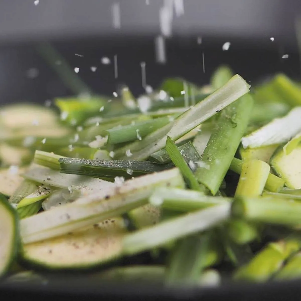 Salt is sprinkled over sliced green zucchini and green onions cooking in a black frying pan.