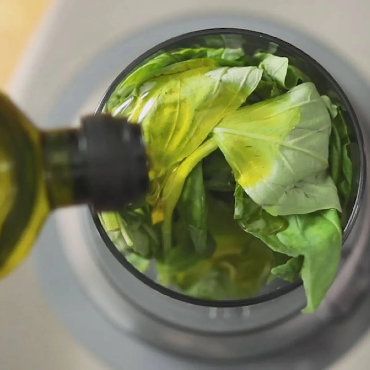 A hand pours olive oil from a green bottle into a food processor filled with fresh green basil leaves.