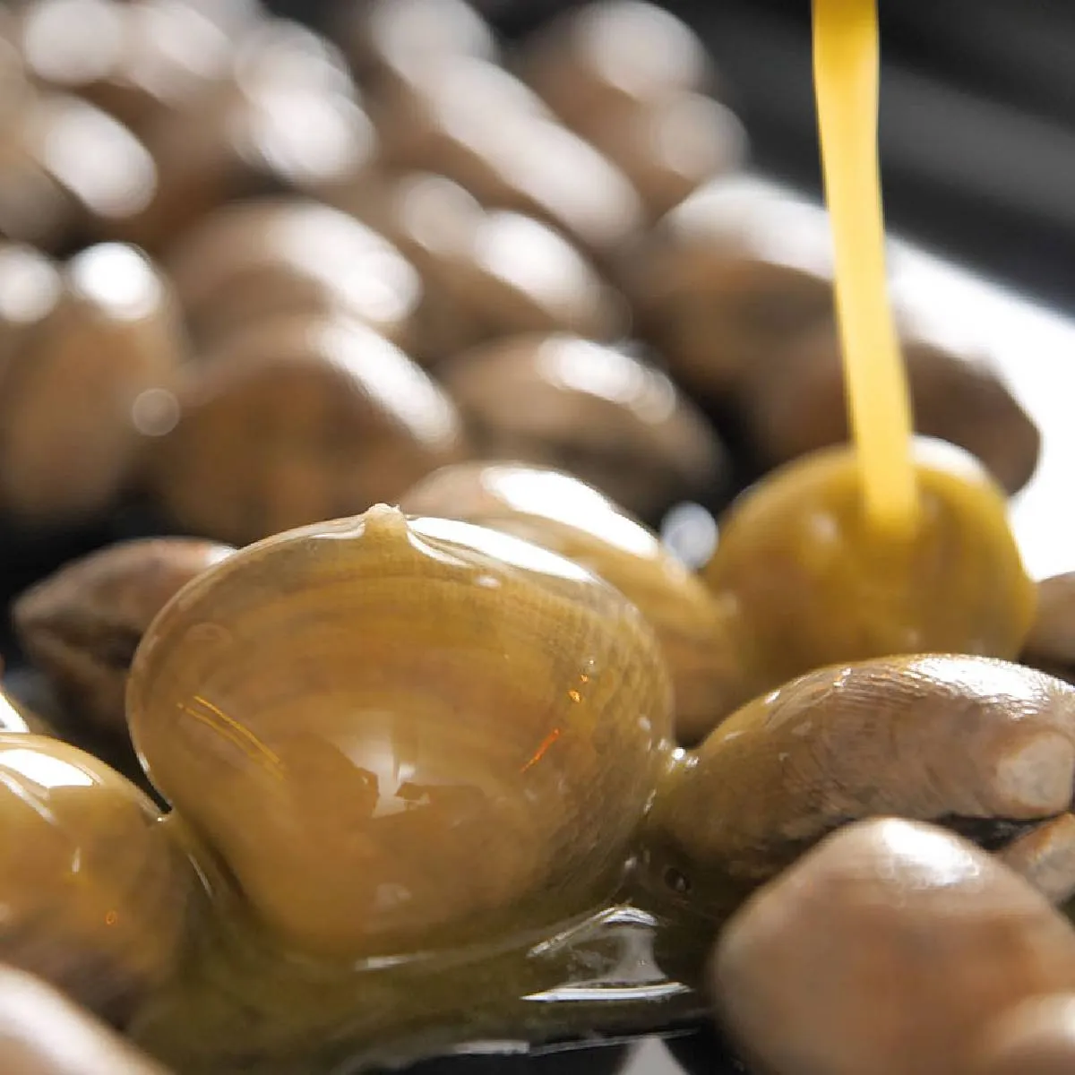 A close-up of a black baking tray filled with clams, being drizzled with a yellow liquid (likely butter or oil).