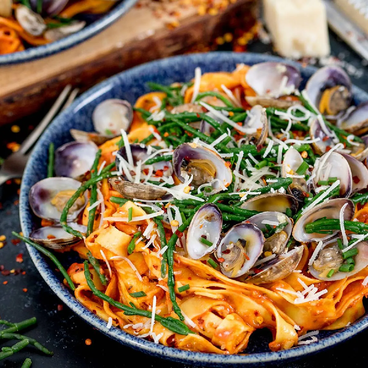 Two bowls of fresh pasta with clams, samphire, and grated cheese, garnished with chili flakes and chives, on a dark blue background.