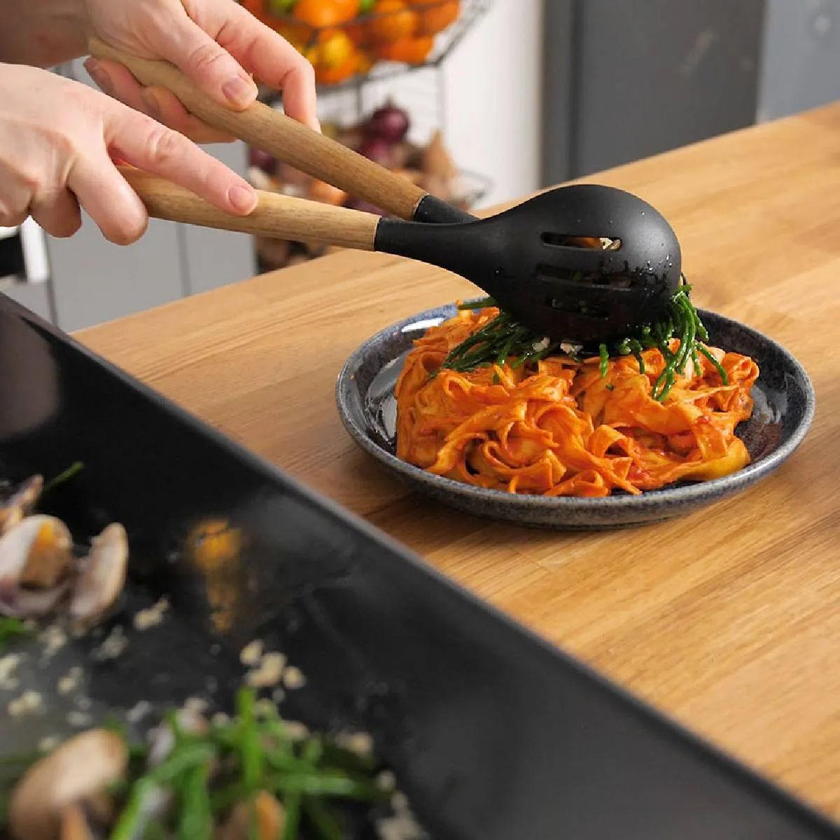 Hands use black tongs to transfer cooked samphire from a black baking tray to a plate of pasta, assembling a dish.