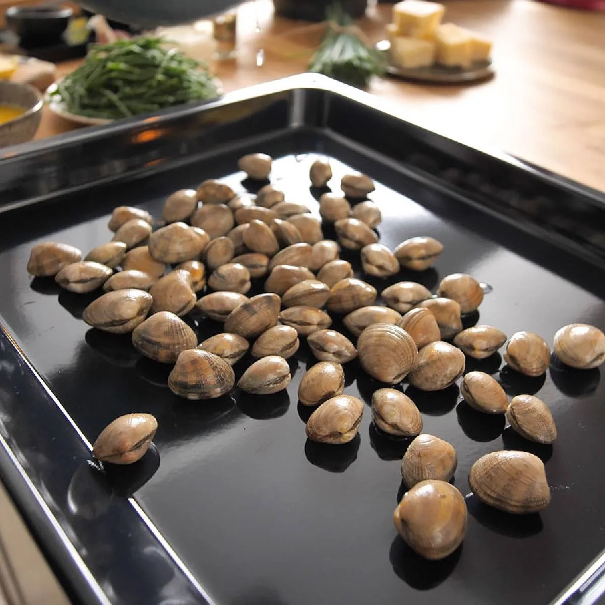 An overhead view of a black baking tray filled with raw clams, ready for cooking, on a wooden countertop.